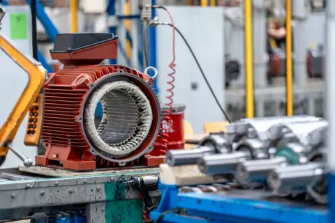 Czerwony silnik elektryczny z widocznym uzwojeniem, przygotowany do montażu. W tle inne rodzaje silników elektrycznych i elementy linii produkcyjnej.