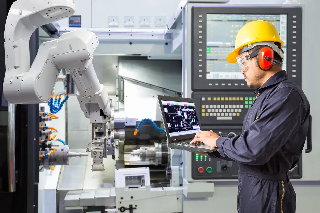 Ciekawostki o robotach: Inżynier w żółtym kasku programuje ramię robota na laptopie, kontrolując nowoczesną maszynę przemysłową.